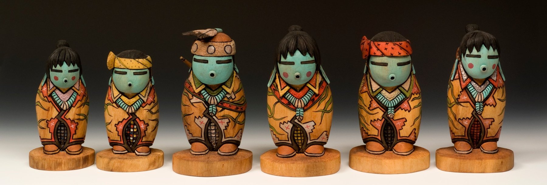 Six carved and painted wooden figures in a row, with turquoise faces, patterned robes, and turquoise necklaces