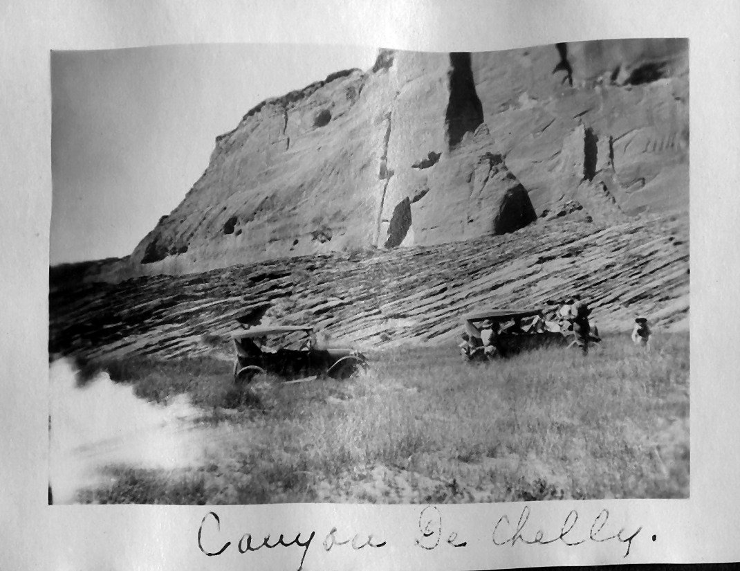 Early automobiles parked on grass below a sandstone cliff in Canyon de Chelly, with a handwritten caption on the photo