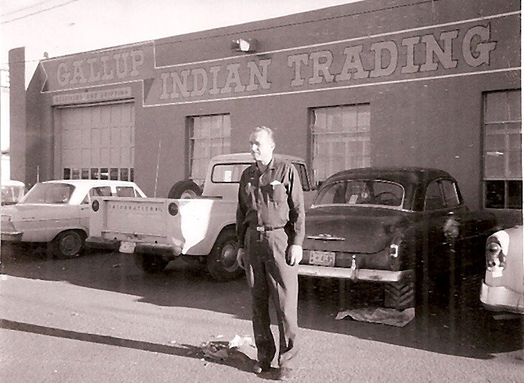 John W. Kennedy standing in front of the Gallup Indian Trading Company building with parked cars and a pickup truck