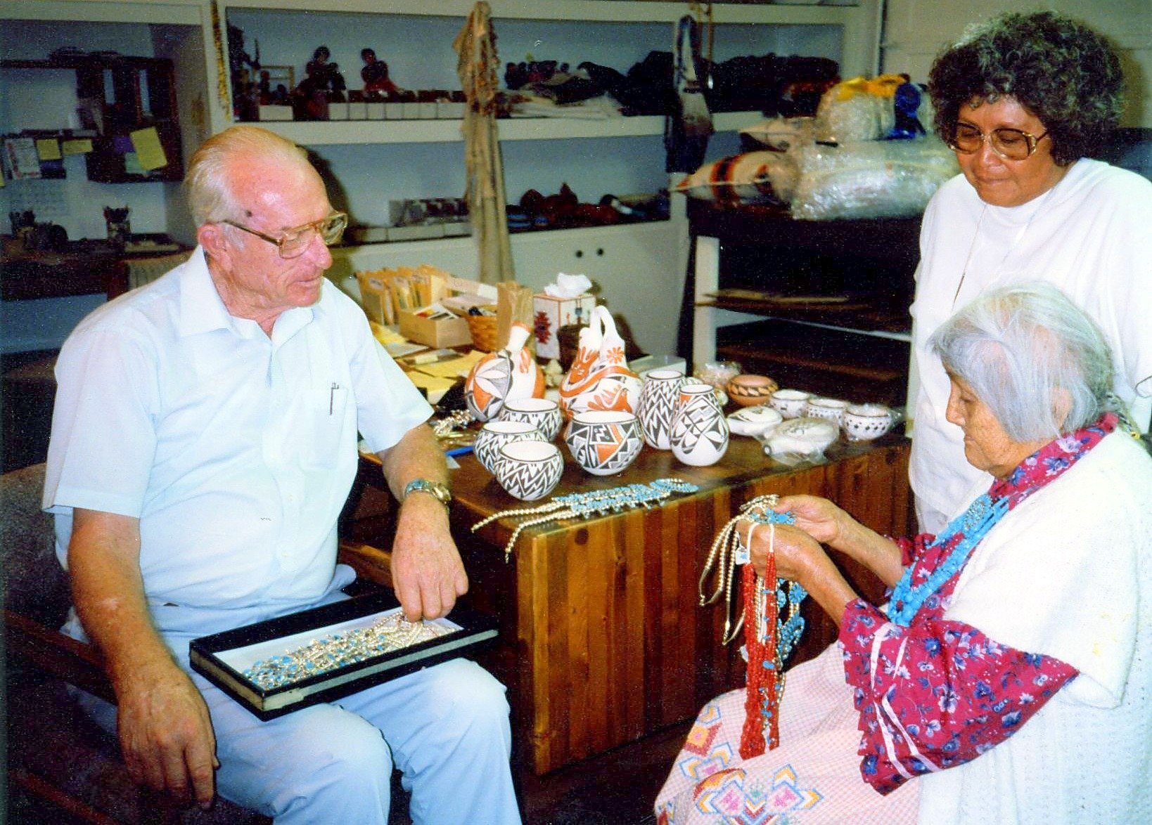 John W. Kennedy seated with a tray of jewelry beside Acoma potter Lucy Lewis and her daughter, with Acoma pottery on the table behind them