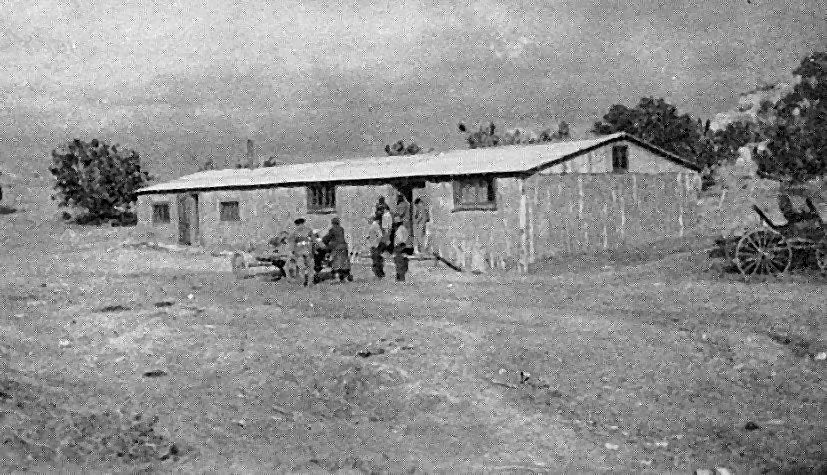 A long, low trading post building with a pitched roof on open ground, people gathered at the door and a horse-drawn wagon at right